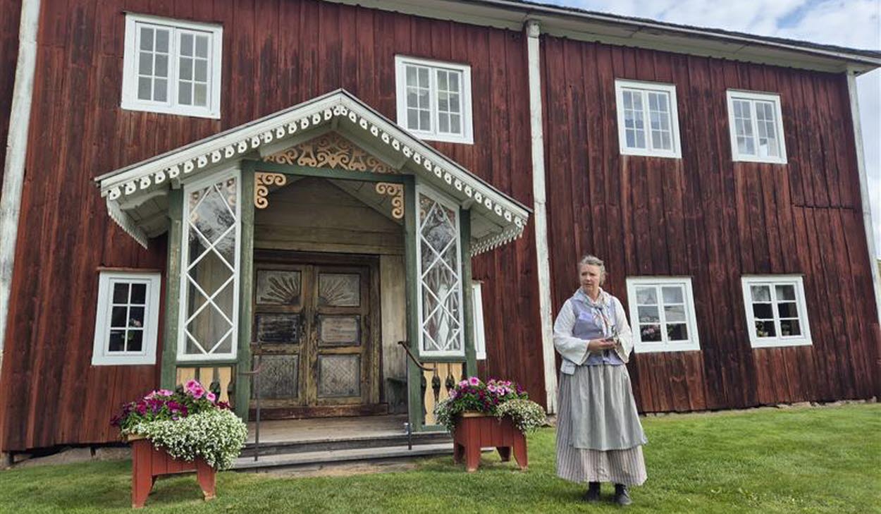 En person i folkdräkt står framför en stor rödmålad träbyggnad med snickarglädje vid entrén.