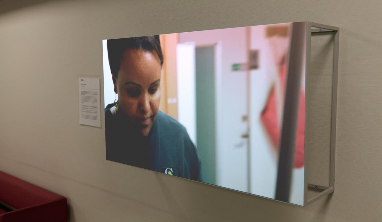 Närbild av skärm på vägg i korridor, som visar en person i arbetskläder i en vårdmiljö.