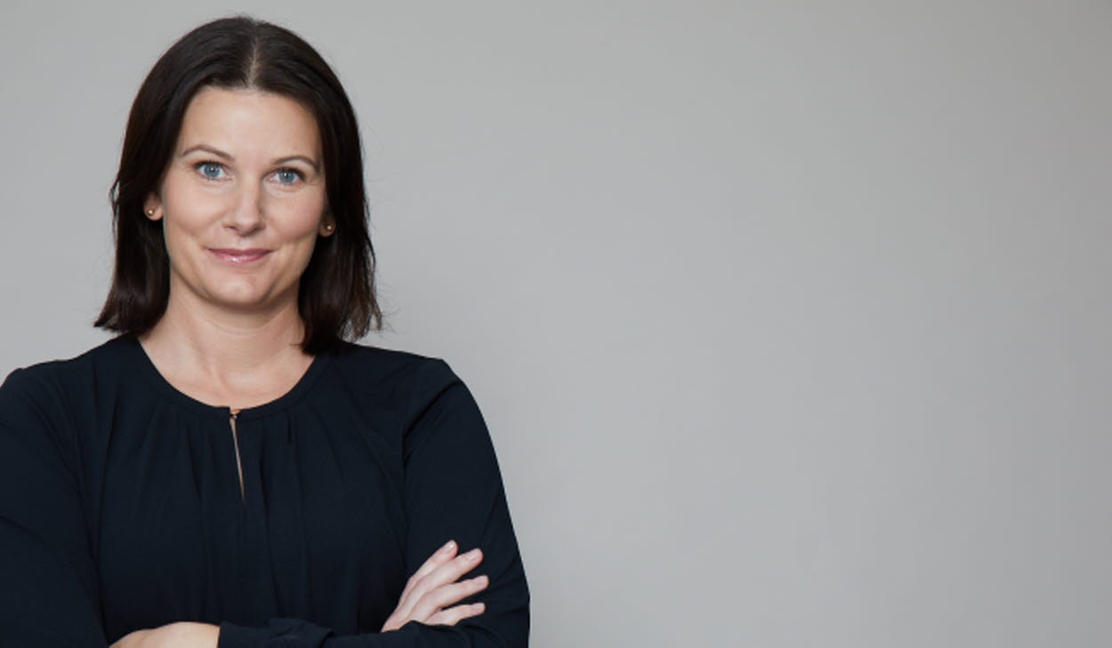 Pressbild på Anna E Wahlgren stående framför en grå vägg, klädd i svart långärmad tröja