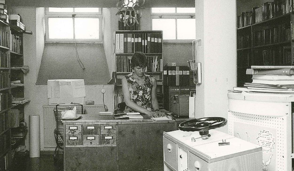 Svartvit bild av ett äldre bibliotek där en kvinna arbetar bakom en trädisk med kortkatalog. Rummet är fullt av bokhyllor, pärmar och dokument i ett trångt men organiserat utrymme.