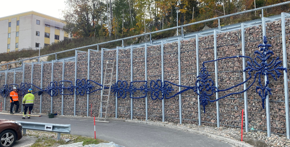 Fyra personer arbetar vid en stödmur där ett blått konstverk monteras mot stenfyllda gallersektioner.