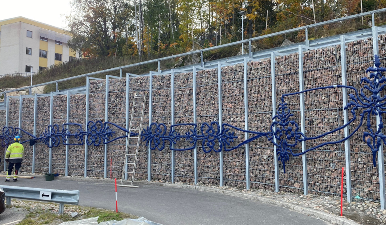 Fyra personer arbetar vid en stödmur där ett blått konstverk monteras mot stenfyllda gallersektioner.